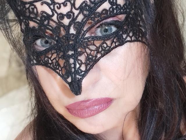 Emerald with pale skin, blue eyes, and dark brown hair, wearing a black lace mask, and pink lipstick, smiling slightly. Her tits are not visible in this shot.
