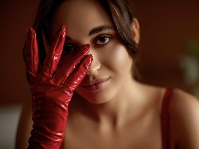 AlexaWilds with shiny red latex gloves, covering one eye, has a subtle smile, brown hair, and a red strap dress, showing off her smooth, sexy face.