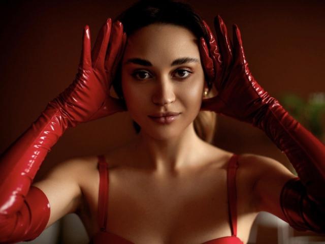 AlexaWilds with red latex gloves, hands on her head, wearing a red dress, brown eyes, slight smile, dark hair in a ponytail, soft brown background.