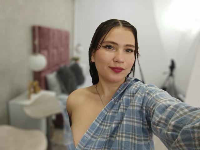 CelesteDash with dark wet hair, plaid shirt, exposing one bare shoulder and small tits, pink lipstick, and black eyeliner, taking a selfie in a bright room.