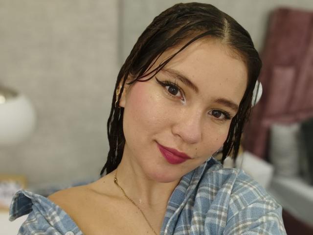 CelesteDash with wet brown hair, pink lipstick, and blue plaid shirt, smiling slightly. She has a gold necklace, and her tits are partially visible. Her face is close to the camera.