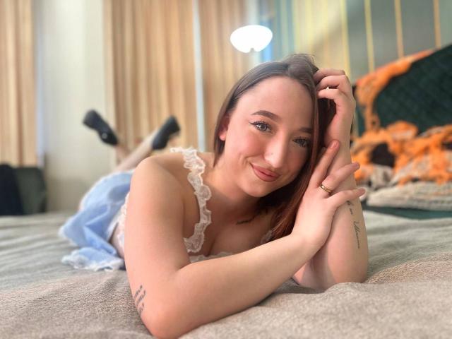 MonAmourR, acostada en la cama, sonriendo, con pechos pequeños, vistiendo una blusa blanca de encaje, jeans azules, zapatos negros, manos cerca del rostro, cabello castaño, tatuaje en el brazo, anillo en el dedo..