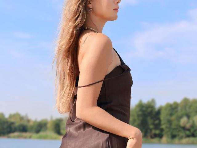 AmmaLoyXX in a brown satin dress with slinky straps, long wavy blonde hair, and a necklace, standing by a blue water body with green trees in the background.