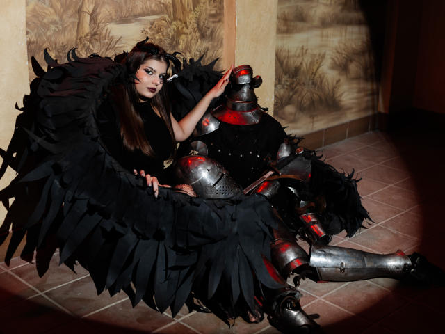 AdeliaLove with black feather wings, sitting on tiled floor, wearing black top and red-accented silver armor, hand on helmet, dark makeup, long brown hair, looking at camera.