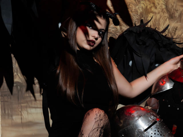 AdeliaLove with dark makeup, black horns, and long brown hair, sitting in front of a metallic helmet, wearing a black top and fishnet stockings, holding black feathered wings.