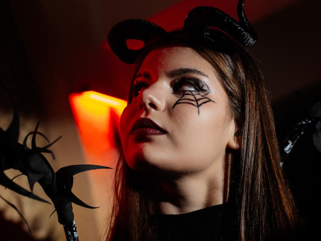 Close-up of AdeliaLove with dark makeup, spiderweb design on her right cheek, black horns on a headband, and straight brown hair. She has a serious expression, slightly upturned face, and dark lipstick. In the foreground, a black, thorn-like object is partially visible on the left. Red-orange light from the background contrasts with her dark makeup and attire. The image has a sharp focus on AdeliaLove's face, with a blurred background.