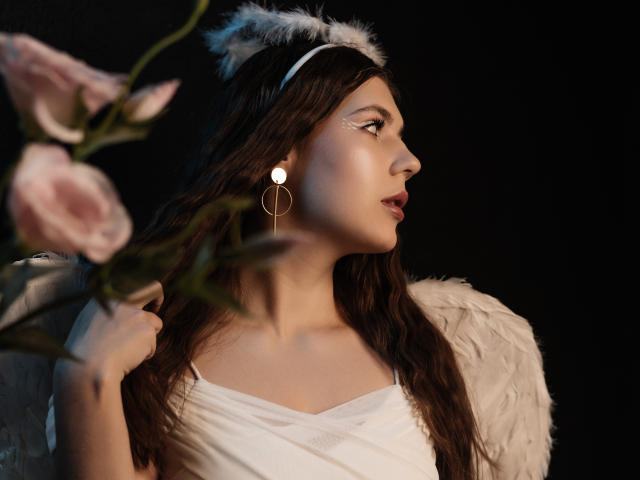 BelleLovely with dark hair, white feather headband, and white dress, holding pink flowers, wearing circular earrings, looking side profile, soft light, dark background.