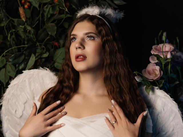 BelleLovely with wavy brown hair, white angel wings, and fluffy halo, hands on her tits, glittery eyes, red lips, and white dress, in a dark setting with flowers.
