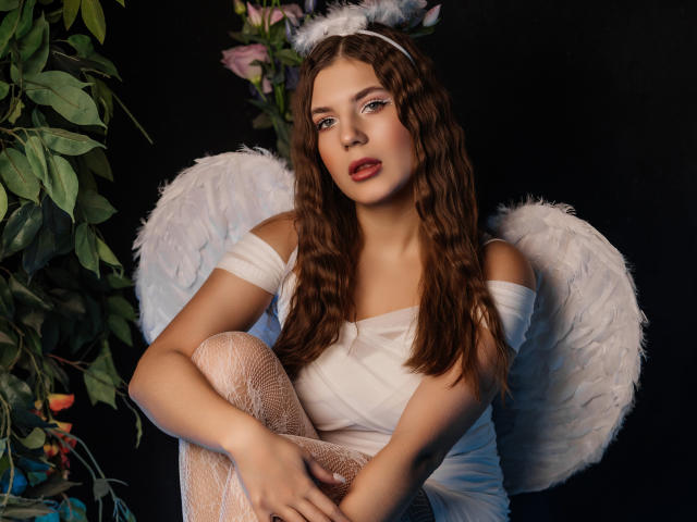 BelleLovely with wavy brown hair, white angel wings, and off-shoulder white dress, sitting, legs crossed, wearing lace stockings, white headband, and red lipstick, against dark background.