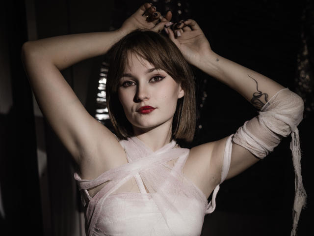 BrittanySweet with pale skin, brown bob, red lipstick, arms raised, wrapped in white bandages, showing small tits, tattoo on right arm, dark background, serious expression, hands above head.