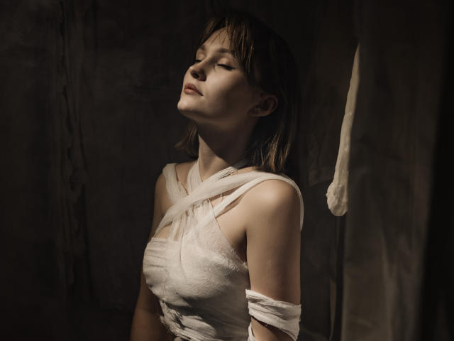BrittanySweet with eyes closed, head tilted back, wearing a white, wrapped-up dress, exposing her shoulders and arms, with soft, dim lighting highlighting her pale skin and brown hair.