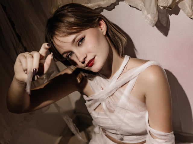 BrittanySweet with short brown hair, red lipstick, and white bandage-like clothing, raising her left hand with black-painted nails, looking at the camera with a slight smile.