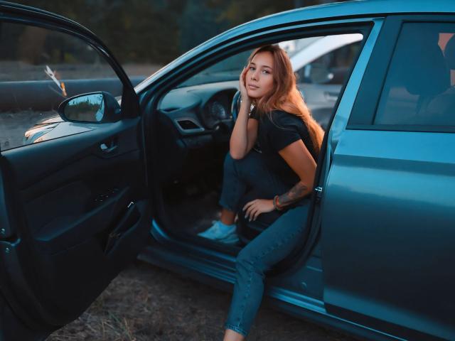 AmmaLoyXX, with long orange hair, sits in a blue car, leaning on the door, wearing a black top and jeans, looking off, tattoo on right forearm, sunset light.