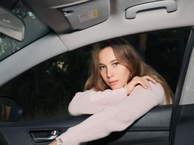 AmmaLoyXX with long brown hair, blue eyes, and pink ribbed sweater leans on car door, arms crossed, looking serious, inside a car with grey headrest and door.