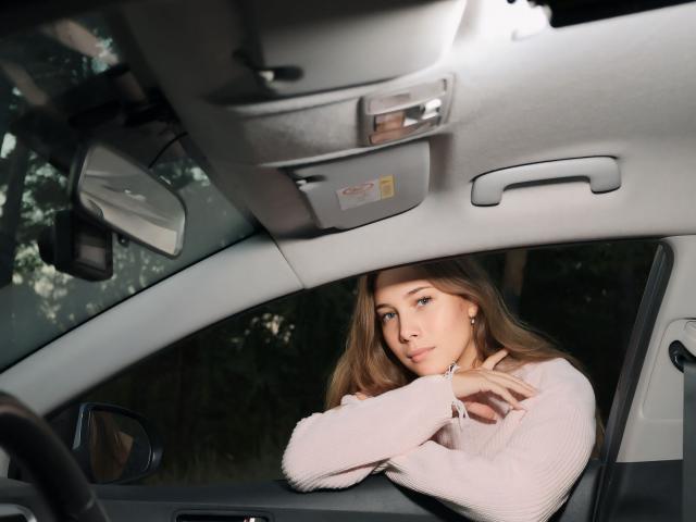 AmmaLoyXX with long blonde hair, blue eyes, and small tits, wearing a pink sweater, leaning on the car's dashboard, looking forward with a serious expression.