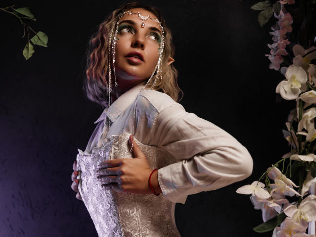 LettieSweet has wavy blonde hair, pointed ears, and wears a bejeweled headpiece. She looks up to the right, her eyes wide. She's dressed in a white lace corset and long-sleeved shirt. She holds a lace-covered book in her right hand. Her left hand is visible, adorned with a red bracelet. White flowers and green leaves are on the right. Dark background. Light from the left. 
   