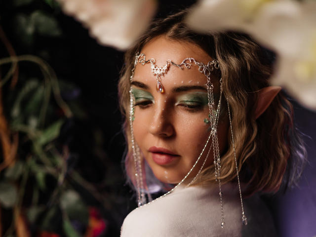 LettieSweet has a fair skin tone and wavy brown hair. She's wearing a delicate silver headpiece with chains and a red gem at the center. Her green eyeshadow contrasts with her pink lipstick. She has a small star tattoo on her right cheek. Her pointed ears suggest an elf-like appearance. She's looking slightly to the side with a calm expression. She's wearing a white garment. The blurred white flowers in the foreground add depth to the image. The dark background is mostly out of focus. The photograph has a soft, almost ethereal quality.