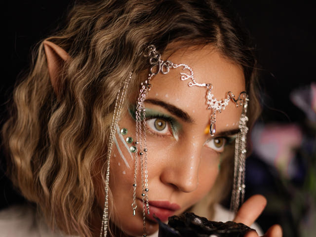 Close-up of LettieSweet with wavy brown hair and elf-like pointed ears. She has green eye makeup with glitter and white speckles on her face. LettieSweet's eyes are light green, looking directly at the camera. She wears a silver metallic headpiece with chains and small beads, covering her forehead. Her right hand, partially visible, holds a dark object near her mouth. The background is blurred, focusing attention on LettieSweet's face. The image has a fantasy or cosplay vibe, with detailed facial makeup and intricate headwear.