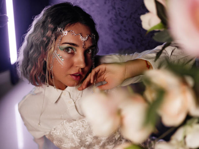 LettieSweet has curly, shoulder-length grayish-purple hair and wears a silver chain headpiece with small ornaments. She has green eyeshadow, dark eyebrows, and a nose piercing. Her white lace dress has a high collar. Her right hand is near her face, touching her cheek. Blurred white and pink flowers are in the foreground. The purple wall is in the background. The image has a soft, warm light.