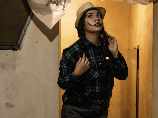MilanaKitty with dark makeup, plaid shirt, brown hat, and suspenders, holding her chin, looking up, in a dimly lit room with yellow walls. Her ass is small.