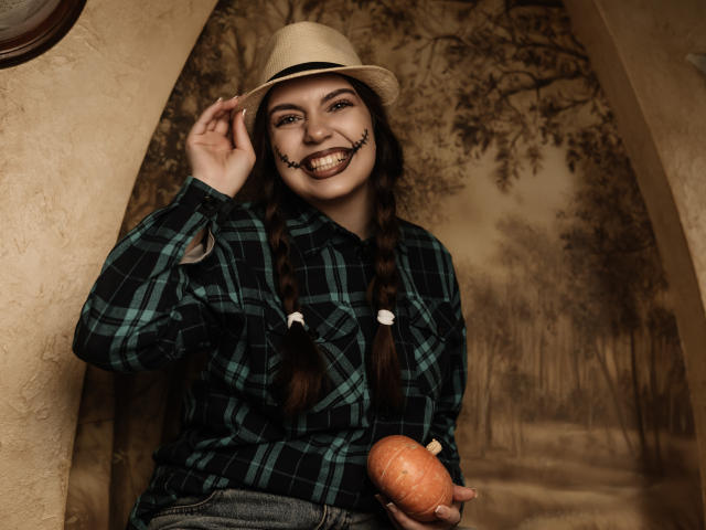 MilanaKitty with pigtails, plaid shirt, and beige hat, grinning with black makeup, holding an orange pumpkin, hand on hat, showing off her cute, small ass.