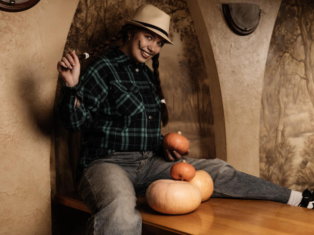MilanaKitty sits on a wooden table with a smile, wearing a green plaid shirt, gray jeans, and a beige hat, holding a pipe, with three pumpkins in front.