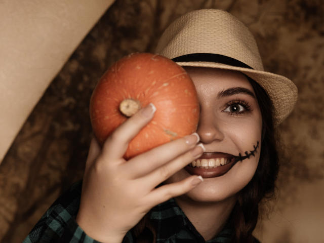 MilanaKitty holds a big orange pumpkin in front of her face, wearing a beige hat and plaid shirt, with dark lipstick and a stitched smile on her cheek.