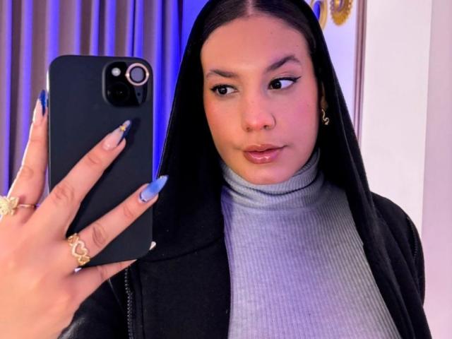 RachellBeaumont taking a selfie, black hair, blue nails, grey turtleneck, black jacket, gold ring, holding black phone, looking at mirror, slight smile, light skin, medium breasts.