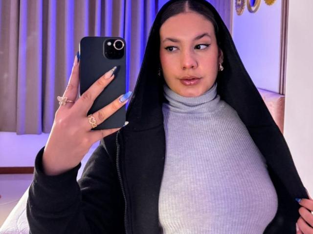 RachellBeaumont with large tits, wearing a black jacket over a gray rib-knit turtleneck, taking a mirror selfie, holding a black phone, long nails, and a confident expression.
