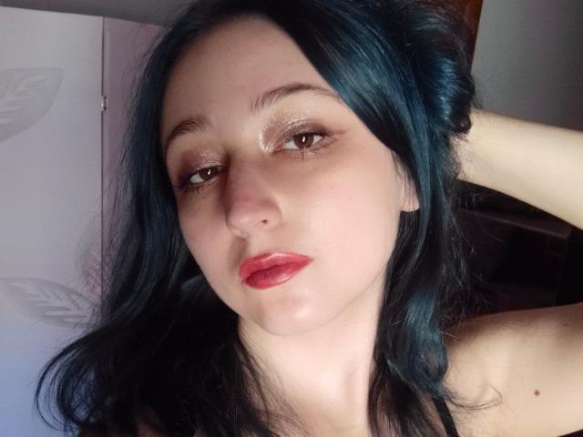 TwinkyKiss has blue hair, shimmering eye makeup, red lipstick, and a slight pout, with a hand in her hair, showing her fair skin and small nose.