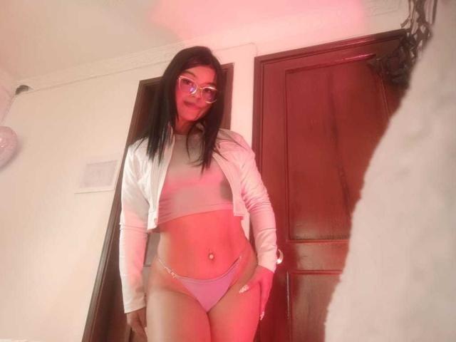 JadeNeferi, con cabello negro largo, gafas, una chaqueta blanca y un bikini rosa, mostrando su abdomen tonificado y su ombligo perforado, está de pie frente a una puerta de madera. Luz roja encendida..