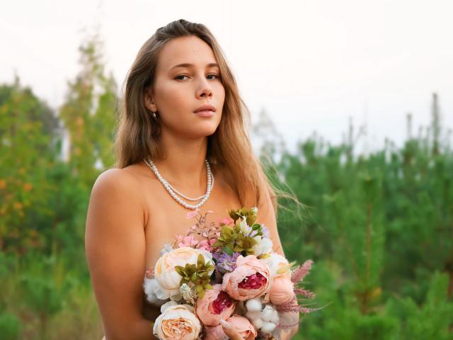 AmmaLoyXX with long brown hair, wearing a pearl necklace, holding a bouquet of pink and white flowers, has a serious expression, showing her bare tits, in a green field.