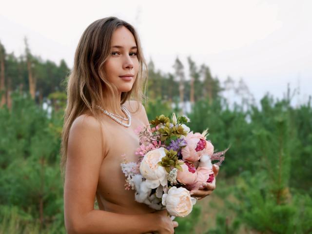 AmmaLoyXX with long brown hair, holding a bouquet of pink and white flowers, wearing a pearl necklace, topless, showing her small tits, standing in a green forest.