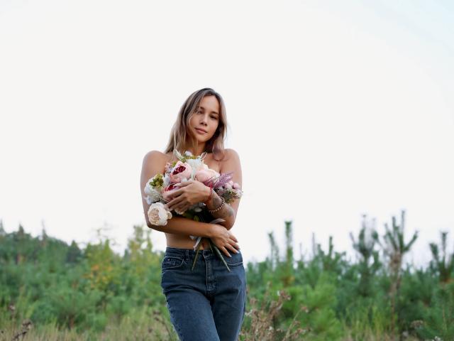 AmmaLoyXX with small tits, wearing a strapless top and blue jeans, holding a bouquet of flowers, standing in a green field, looking slightly sad.