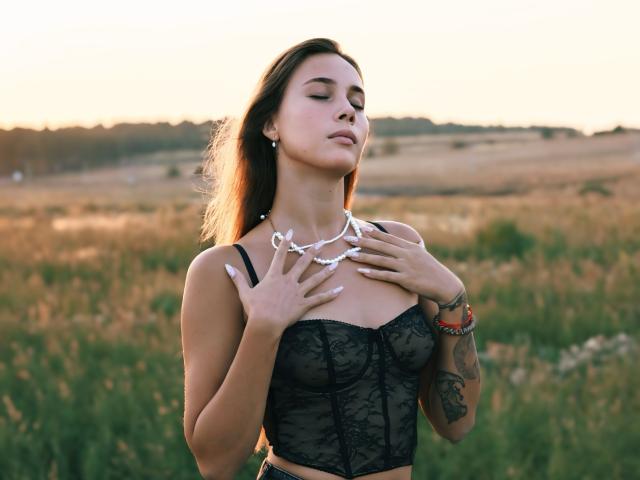 AmmaLoyXX with long brown hair, eyes closed, wearing black lace corset, white necklace, hands on chest, tattoo on right arm, standing in grassy field at sunset.