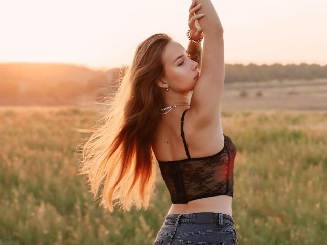 AmmaLoyXX with long brown hair, black lace bra, and blue jeans, raising arms, sunlight, field, soft focus, slight wind, cute, attractive, small tits, slim, standing.