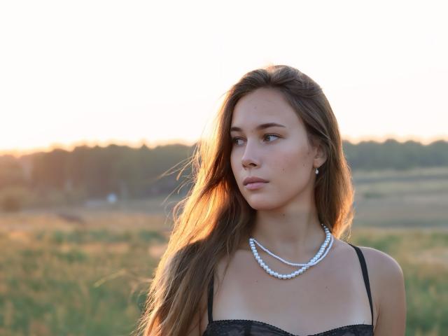 AmmaLoyXX has long brown hair, wearing a black strap dress with a pearl necklace, looking to the side with a serious expression. Her tits are small, in the shot.