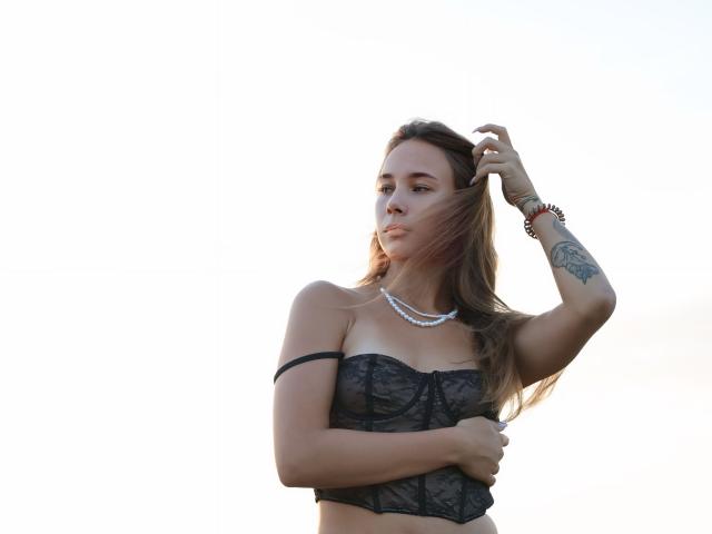 AmmaLoyXX with long brown hair, wearing a black lace bustier, pearl necklace, and bracelet, touching her hair, showing a tattoo on her right forearm, and holding her tits.