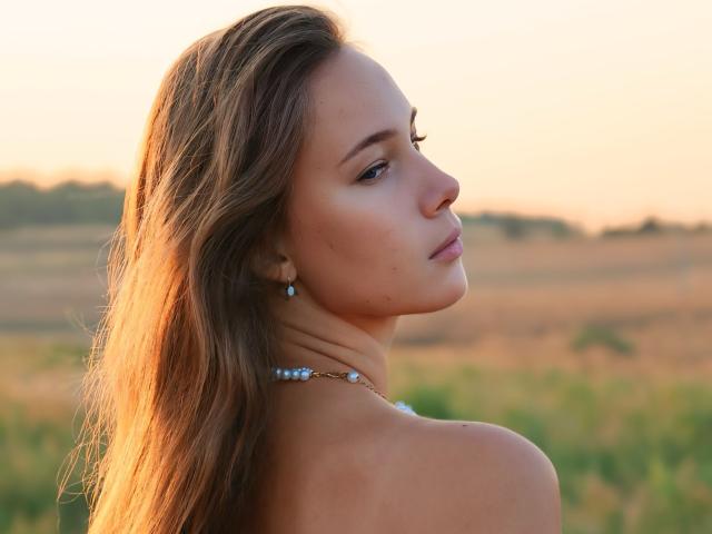 AmmaLoyXX with long brown hair, blue earrings, and a necklace, looking over her shoulder, showing her bare shoulders and soft, pretty face, in natural light, with a calm expression.