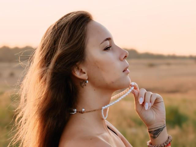 AmmaLoyXX with long brown hair, holding a string of white beads to her mouth, showing a tattoo on her right wrist, in a sunlit, grassy field.