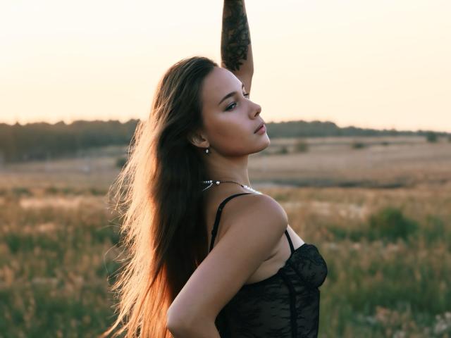 AmmaLoyXX with long brown hair, wearing black lace lingerie, raising tattooed arm, looking back with pouty lips, sunlit field in background, soft golden light, sexy pose.