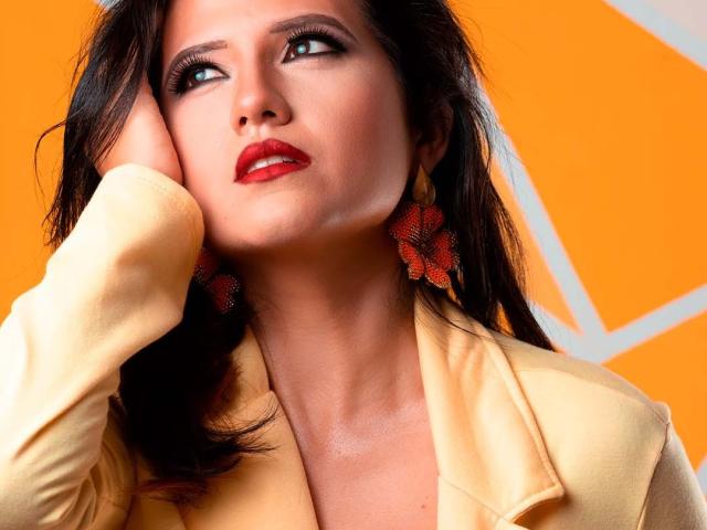 Renata Rivas, met rode lippenstift, donker haar, een gele jas, met haar hand op haar wang, naar boven kijkend, draagt rode bloemoorringen en heeft een sexy, verleidelijke uitdrukking. Haar borsten zijn licht zichtbaar..