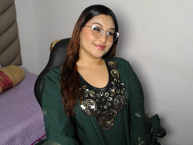 HelueJamil has brown hair, glasses, and a green dress with black, swirling, ornate patterns on the chest. She's sitting, smiling, and looking at the camera.