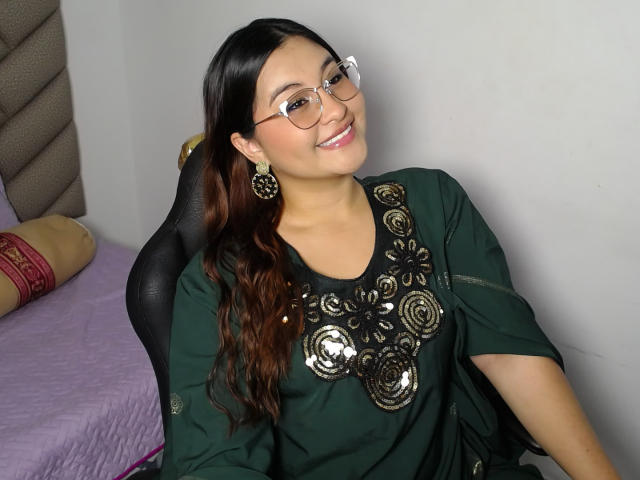 HelueJamil with brown hair, glasses, and dark green top with shiny floral patterns, smiling, sitting, wearing large floral earrings, on a black chair, in a room.