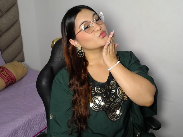 HelueJamil with glasses, long brown hair, green shirt with gold patterns, touching cheek, wearing earrings and bracelet, sitting in black chair, pink bed in background.