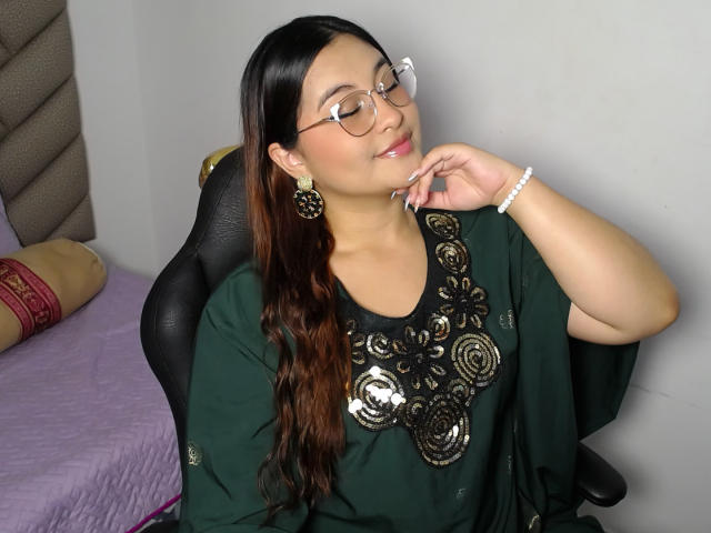 HelueJamil, con lunghi capelli castani, occhiali, una maglia verde con motivi floreali dorati, sorridente, che si tocca il mento, indossa orecchini verdi e un braccialetto bianco, seduta su una sedia nera..