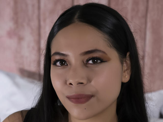 AnnieHill with black hair, brown eyes, and pink lipstick, smiling subtly, small mole below her upper lip, wearing small gold earrings, looking at camera.