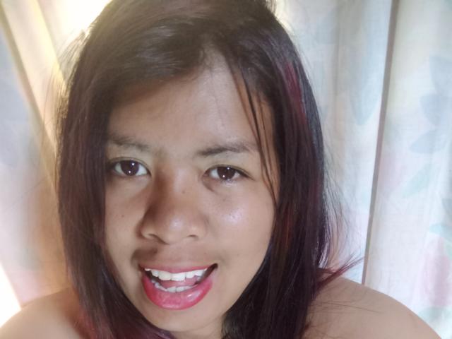 MariahSquirting69 has medium brown skin, dark brown eyes, and a smiling face with pink lipstick. She has straight, dark brown hair with red highlights. Her nose is small.