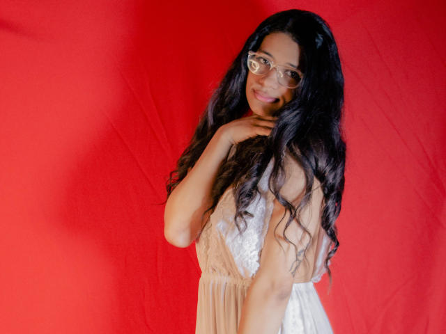 BonnyEbonny, com longos cabelos negros ondulados, óculos e um vestido bege e branco, sorrindo suavemente, tocando o pescoço, em pé contra um fundo vermelho brilhante..