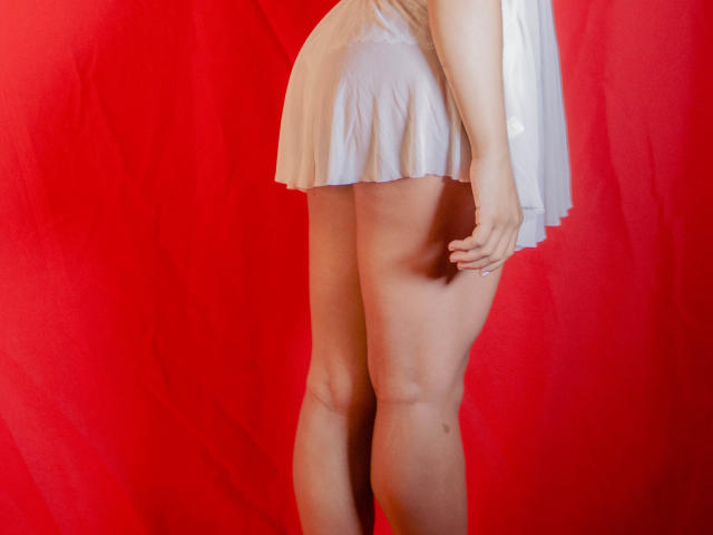 A photograph of BonnyEbonny from the back, showing her lower body and legs. She's wearing a short, white, sheer dress that flares slightly. Her right hand is partially visible, resting near her thigh. The background is a solid red, creating a stark contrast with her light skin and the white dress. Her legs are slender and smooth, with a slight shadow on her right thigh. The composition focuses on the lower half of her body, emphasizing her legs and the dress's texture.